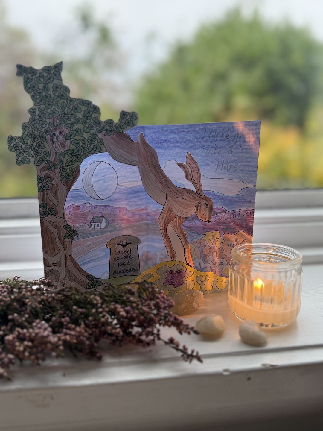Handmade paper scene of Isobel Gowdie as a hare under the Seed Moon, with a candle and heather on a windowsill.